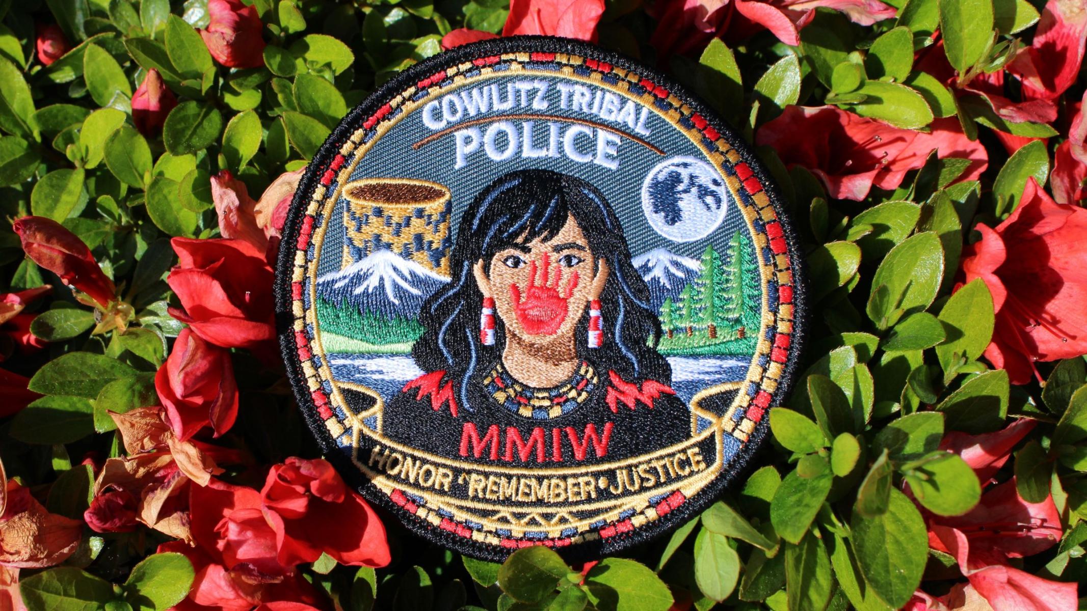 Red flowers surrounding a patch stating Cowlitz Tribal Police, showing a Native woman with red hand covering her mouth. Below states MMIW and Honor, Remember, and Justice.