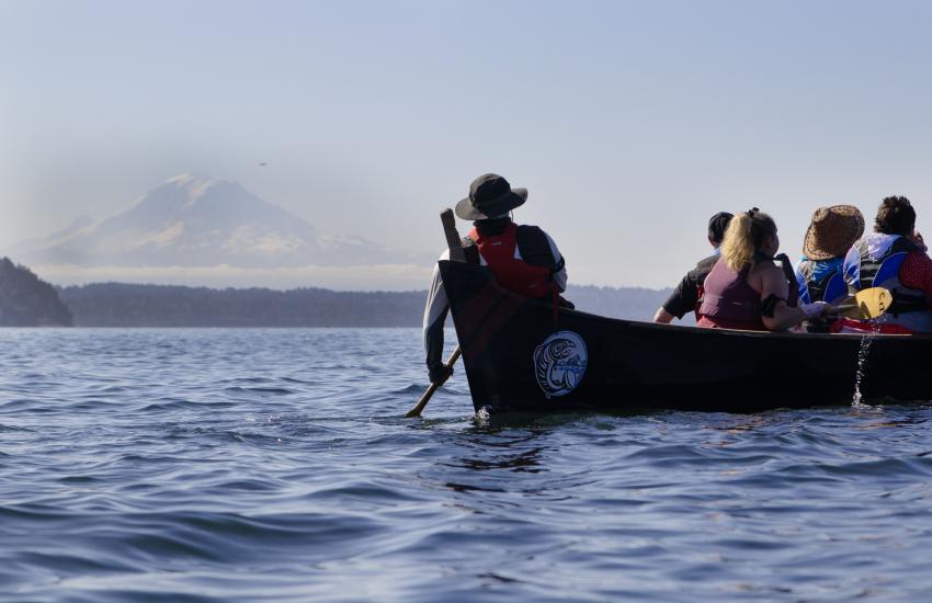 Cowlitz Canoe Family paddles across the water in a traditional canoe, with Mount Rainier visible in the distance.