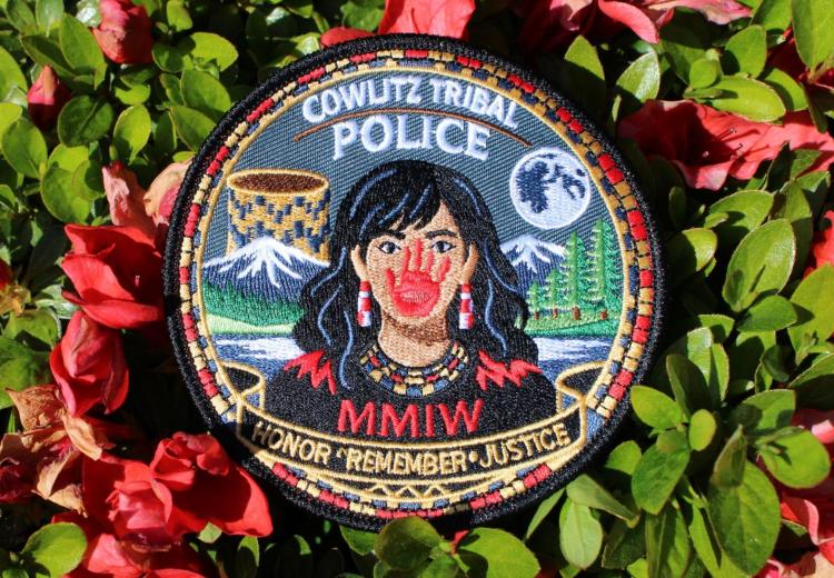 Red flowers surrounding a patch stating Cowlitz Tribal Police, showing a Native woman with red hand covering her mouth. Below states MMIW and Honor, Remember, and Justice.